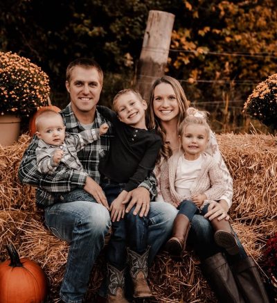 Picture of two people holding three kids. They are all smiling and sitting on hay bales surrounded by pumpkins and pots of flowers.