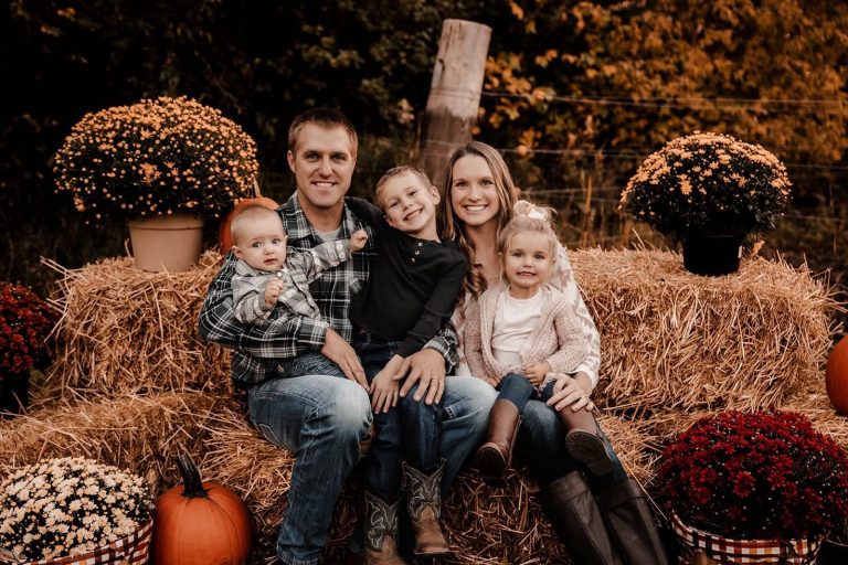 Picture of two people holding three kids. They are all smiling and sitting on hay bales surrounded by pumpkins and pots of flowers.