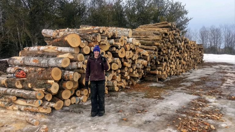 A woman standing in front of a large stack of lumber for firewood.