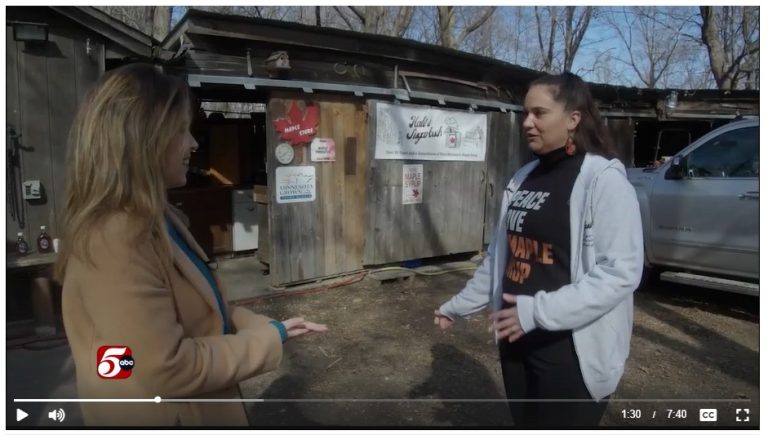 A woman speaks to a reporter in front of a maple syrup farm stand.