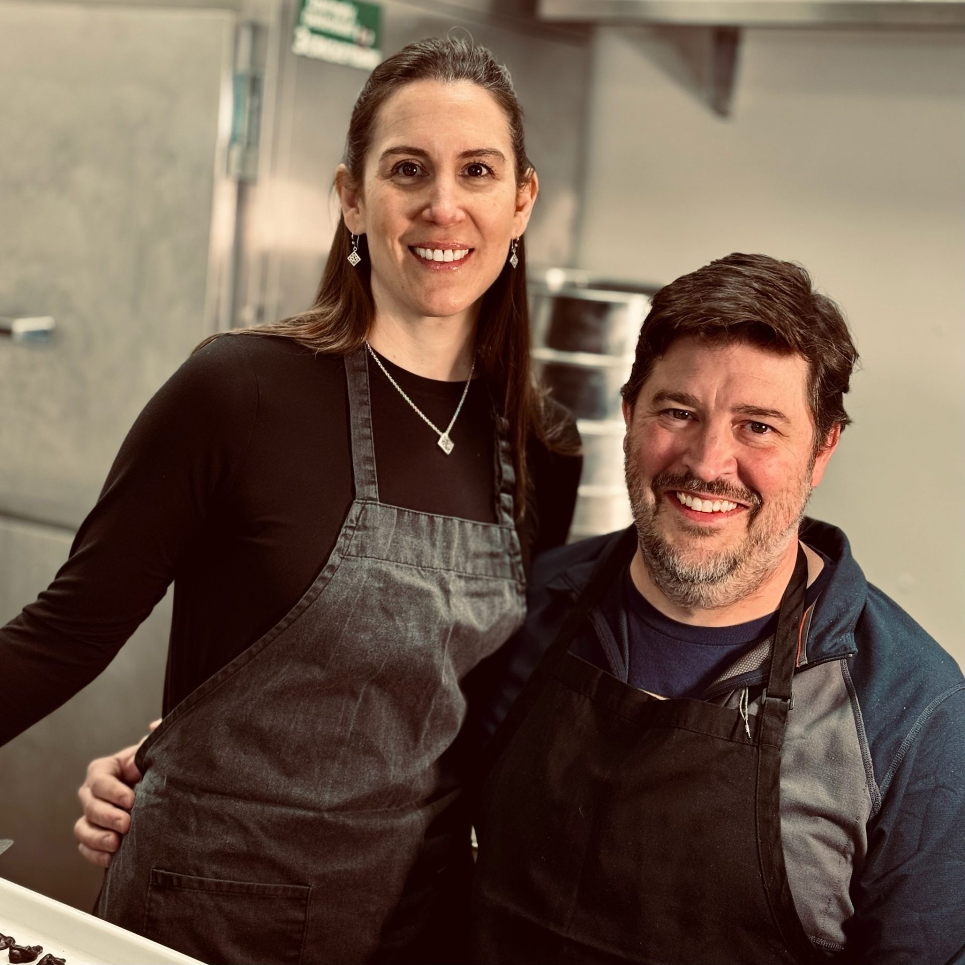 Two people with kitchen aprons on smiling in what looks to be a commercial kitchen.