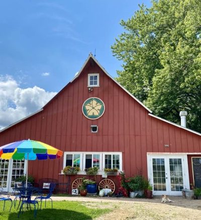 Picture of red barn in the sunshine with a blue sky and a table with a rainbow umbrella in the front.