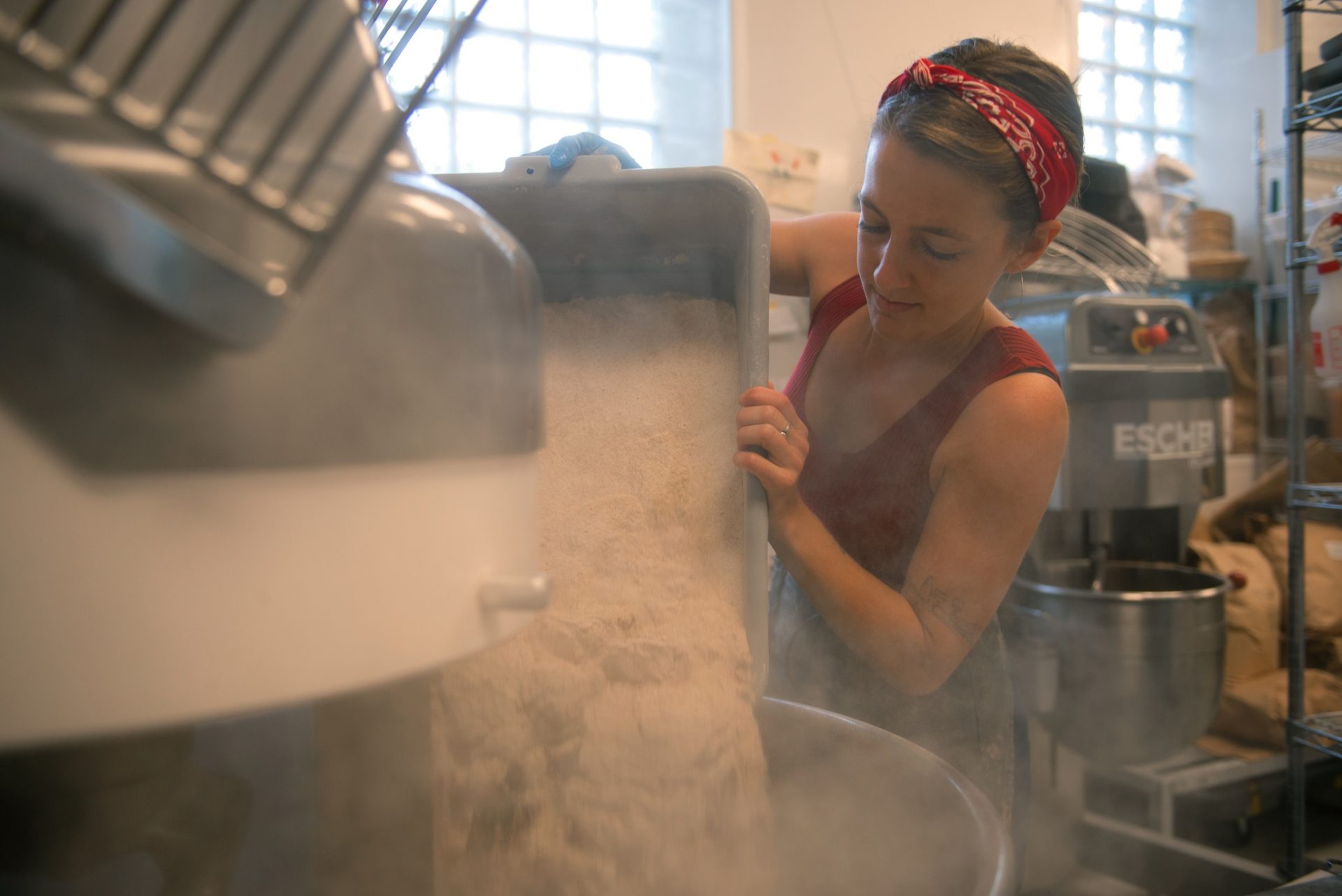 A woman pouring flour into a large industrial mixer.