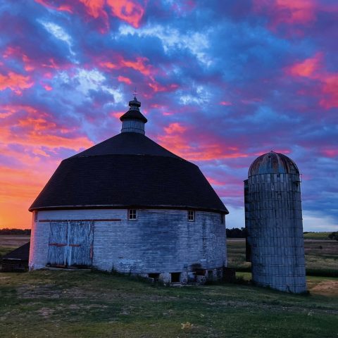 A round white barn and silo with a pink and blue sunset behind it.