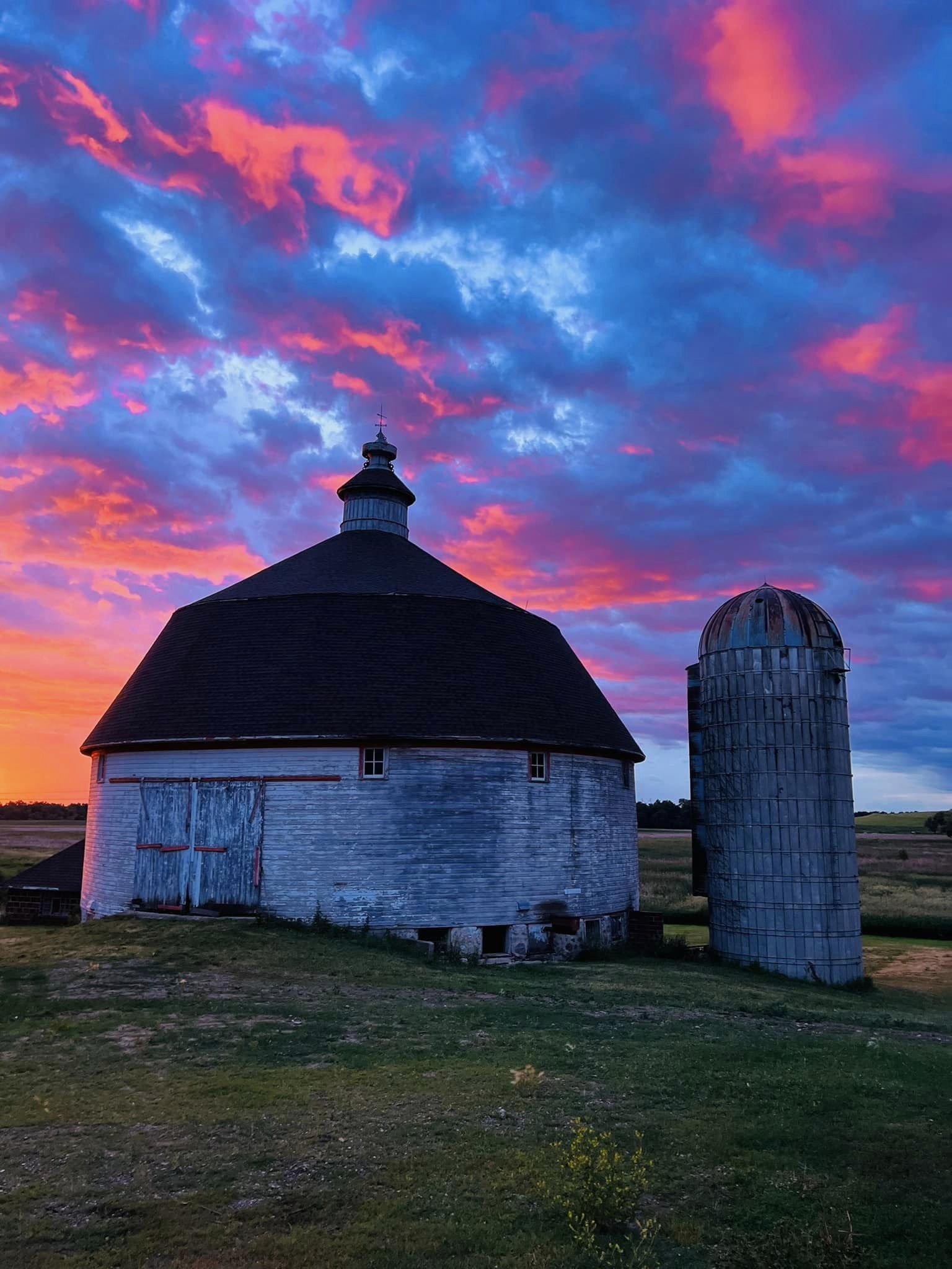 A round white barn and silo with a pink and blue sunset behind it.