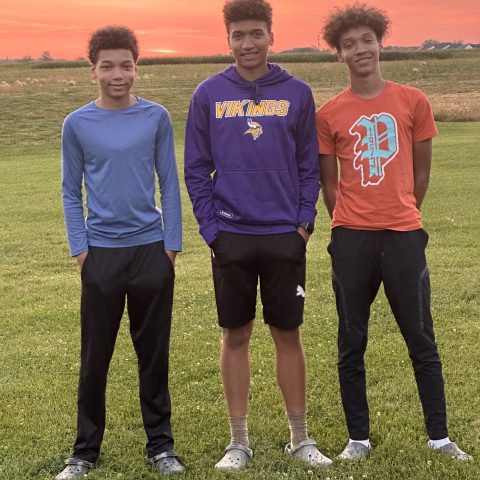 Three teenage boys stand in a field with a sunset behind them.