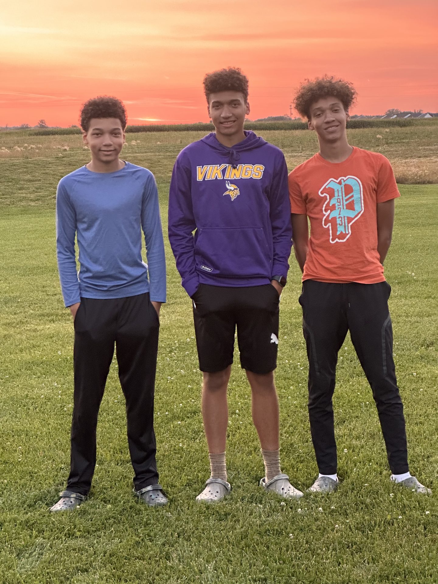 Three teenage boys stand in a field with a sunset behind them.