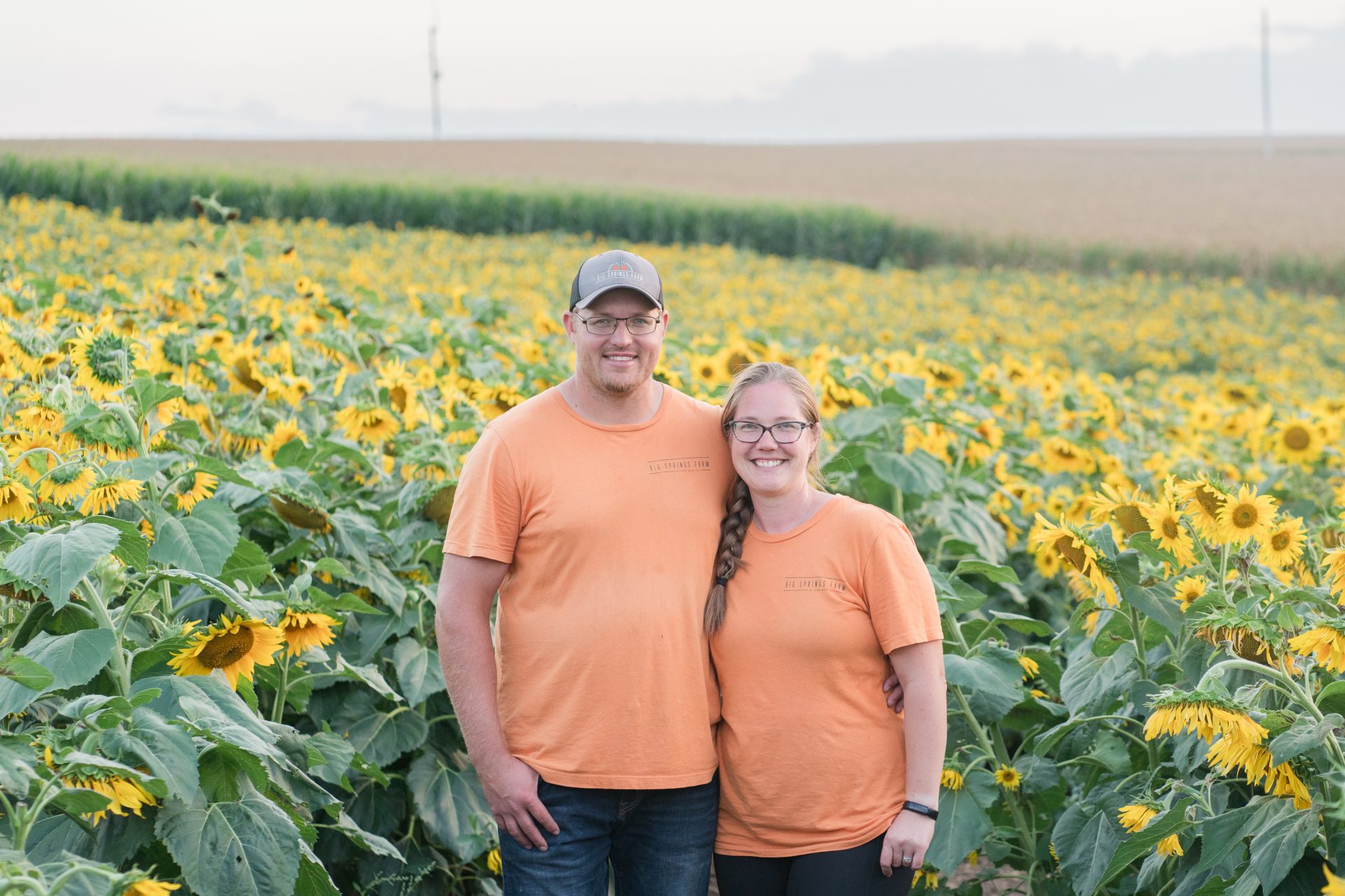 Two farmers, a man and a woman in matching orange t-shirts, standing in a field of sunflowers.