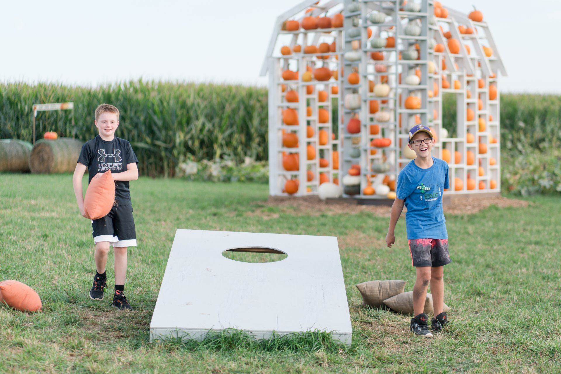 Two boys playing bean bag toss in front of a structure filled with pumpkins.