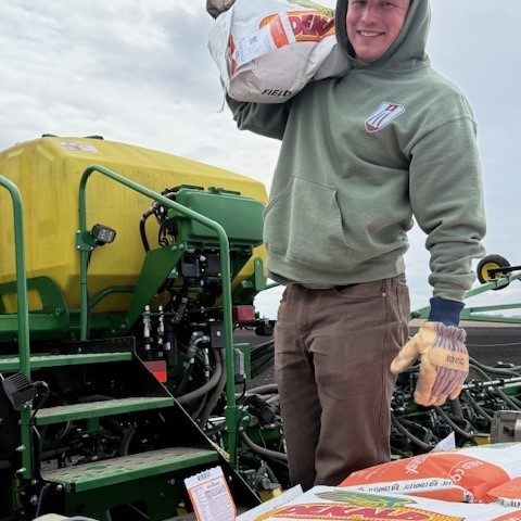 young man hold bag of seed over his shoulder and smiles, tractor is behind him