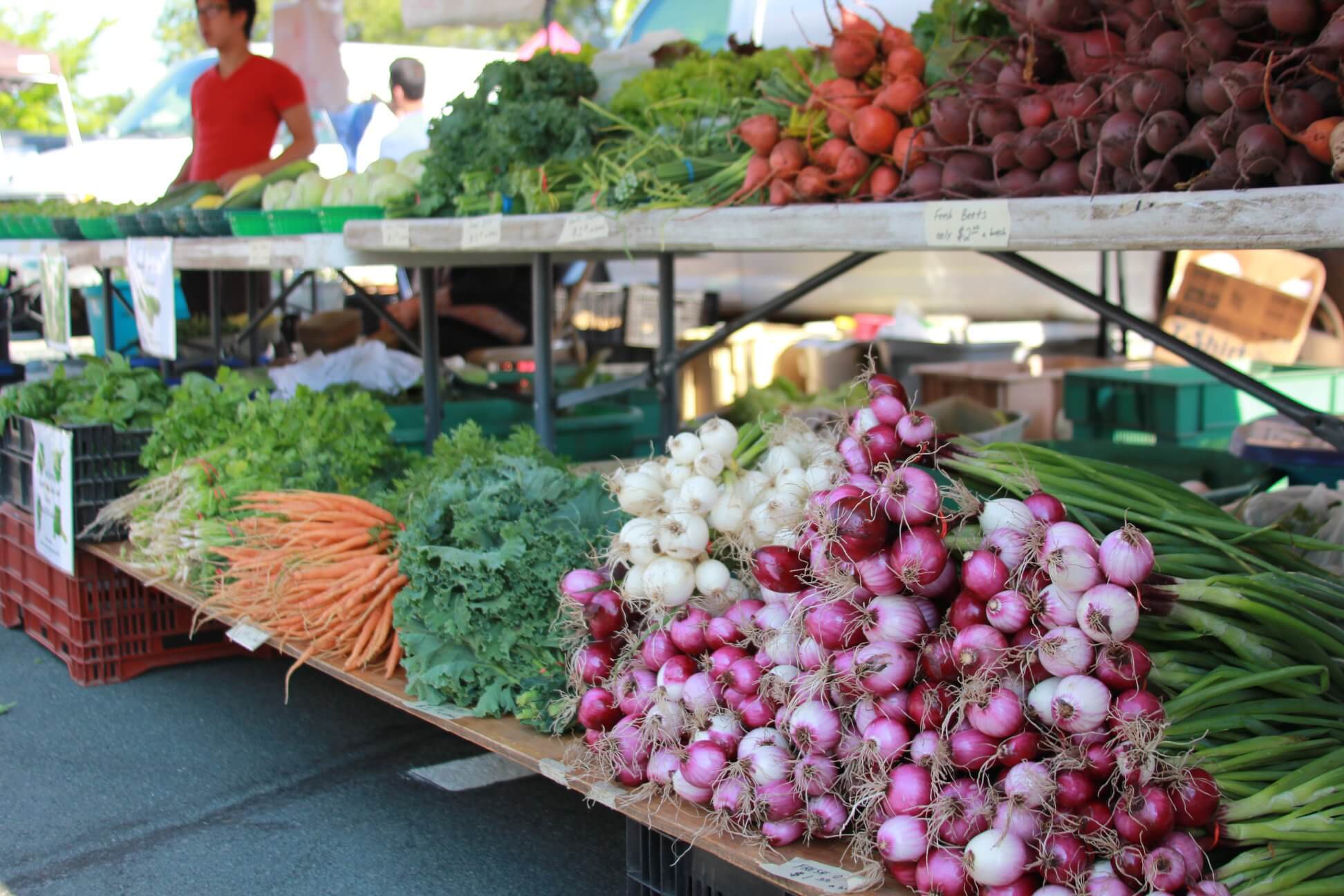 Picture of shelves of onions, carrots, cilantro, beets, radishes, broccoli, and other vegetables at a farmers' market