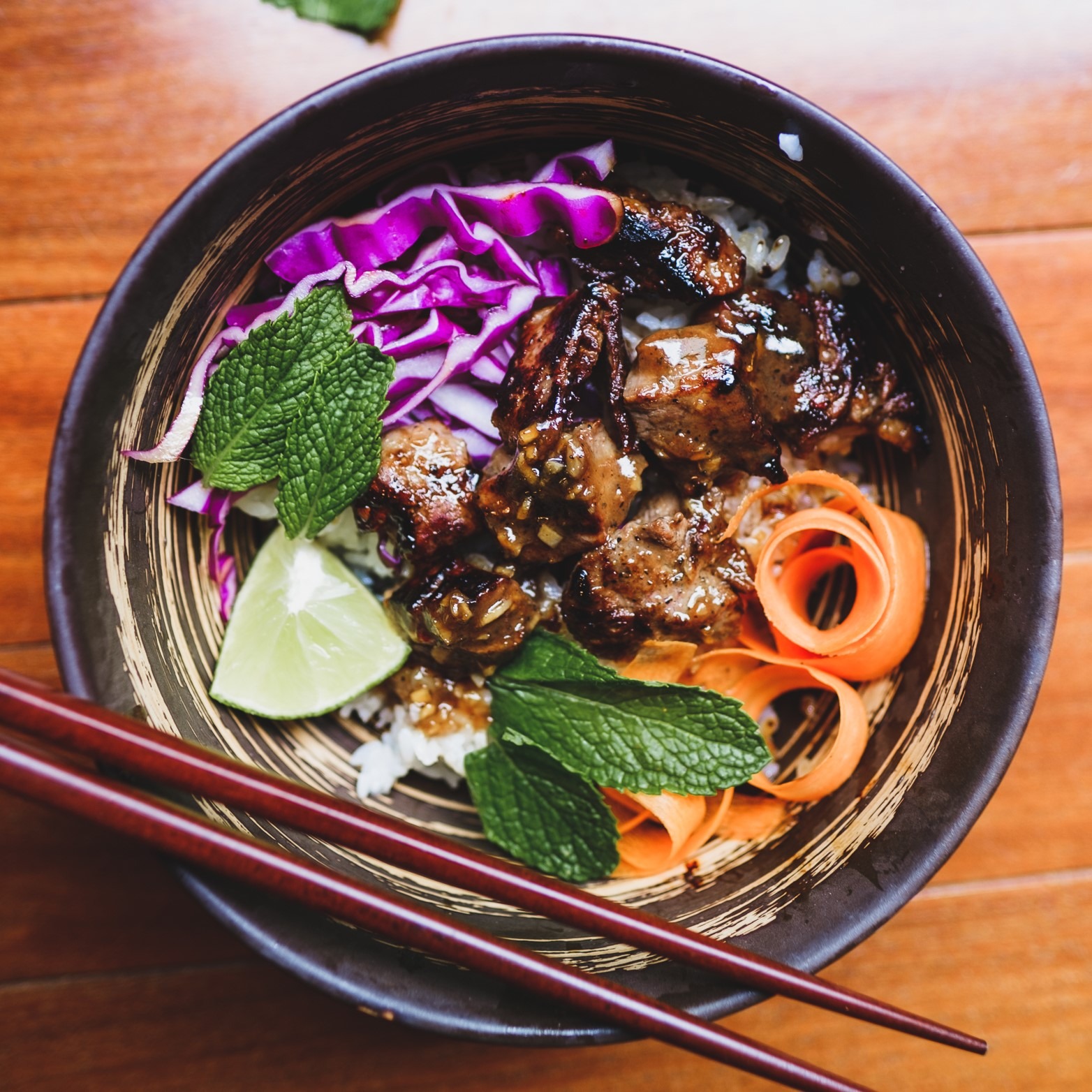 Picture of a rice bowl with purple cabbage, meat coated in sauce, carrots, mint leaves, and lime. A set of chopsticks sits on the edge of the bowl.