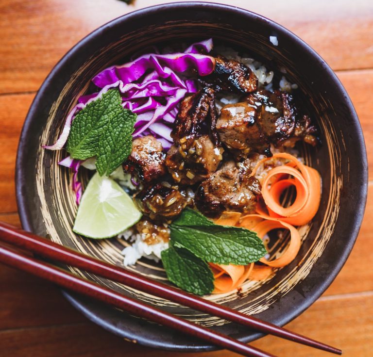 Picture of a rice bowl with purple cabbage, meat coated in sauce, carrots, mint leaves, and lime. A set of chopsticks sits on the edge of the bowl.