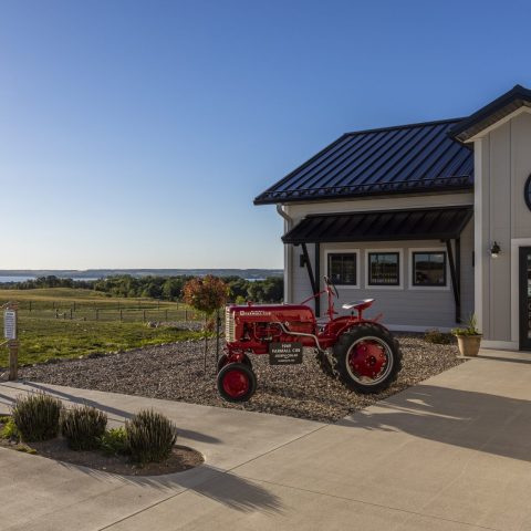 A picture of a red tractor parked in front of a building with the vineyard in the background.