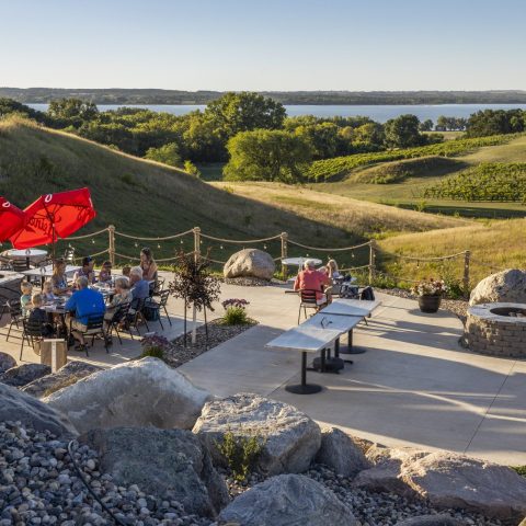 A picture of people sitting at tables on a patio outside with rolling hills of vineyards in the background.