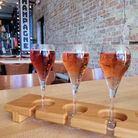 three glasses of rose sit in a flight holder. a large sign in the background reads CHAMPAGNE