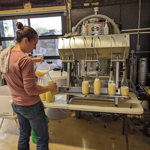a woman bottles wine, her back facing the camera. the bottle on the table to her right is promenent.