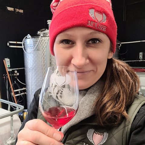 a woman smiles, she holds a glass of rose. the glass, her pink beanie hat, and her gray vest all feature the mousse logo