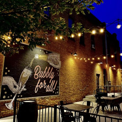 patio at night with string lights up above, mural reads BUBBLY FOR ALL and showcases a bottle pouriing into a glass