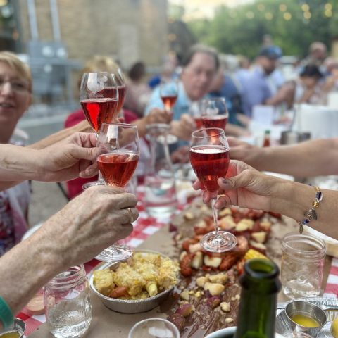 a full table surrounded by people blurred in the background, hands gather to cheers wine glasses filled with rose