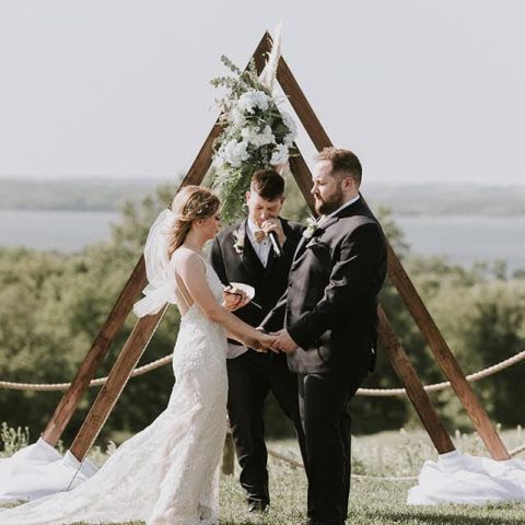 A bride and groom stands at a wedding alter in the vineyard, holding hands.