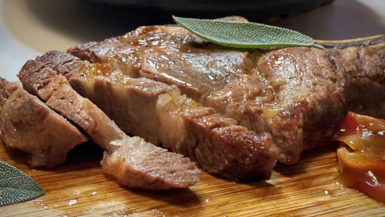 Photograph of a pork chop on a cutting board. It is topped with a sage leaf and there are baked apples on the side.