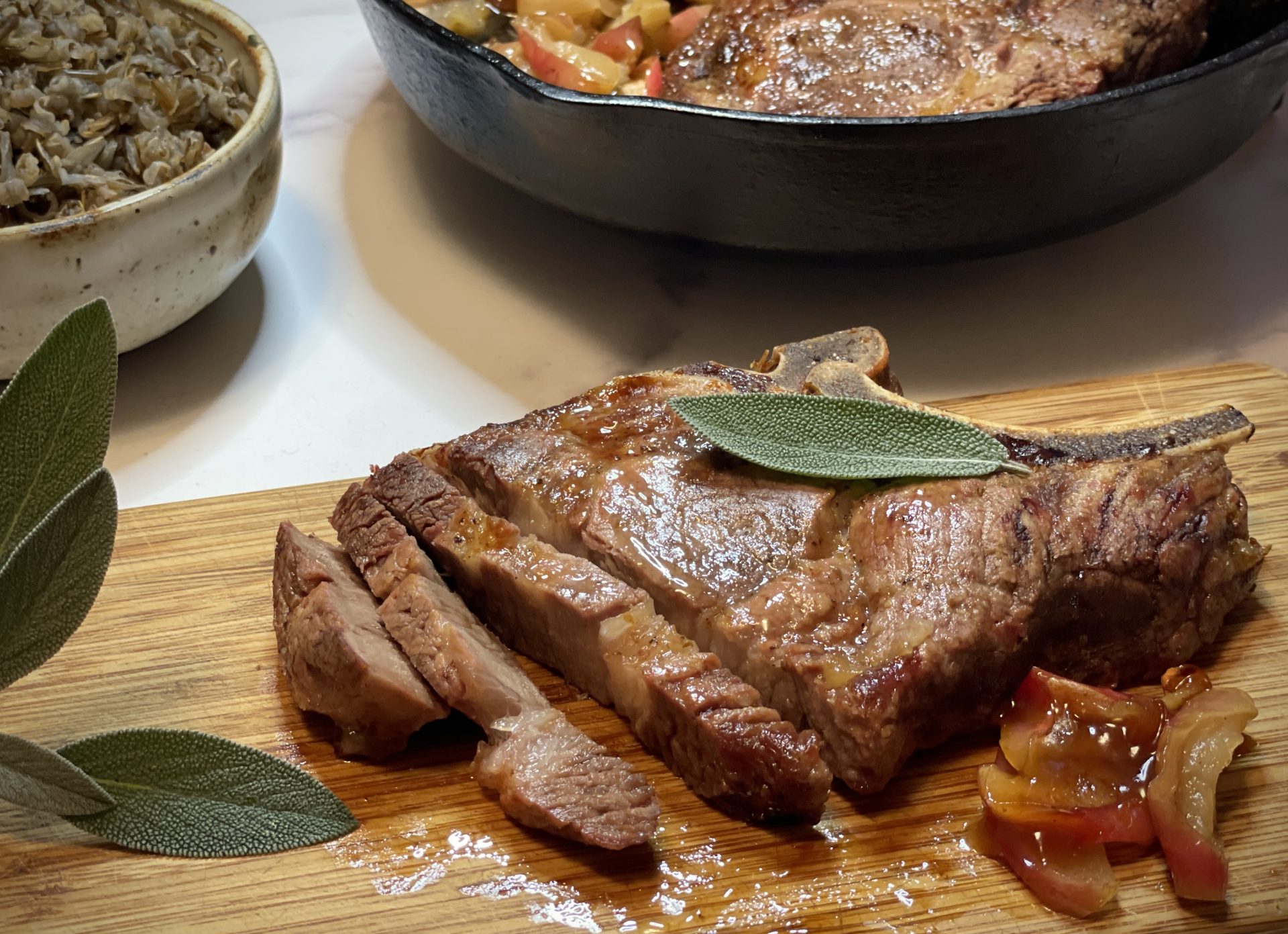 A pork chop on a cutting board. In the background is a bowl of rice and a cast iron pan with pork and apples in it.