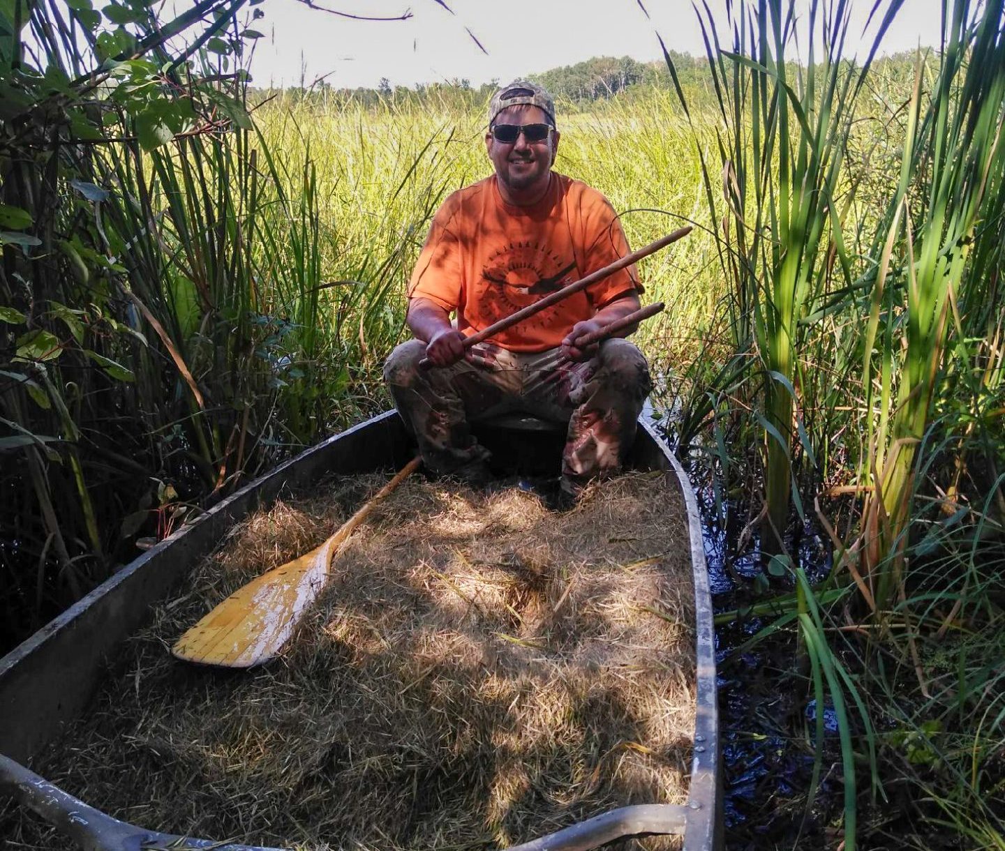 Smiling person sitting in canoe filled with wild rice grains, with wild rice plants on both sides of the canoe and a lake in the background