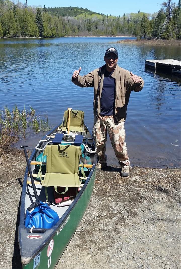 Person standing next to canoe on shore of a lake. Person is smiling and giving double thumbs up.