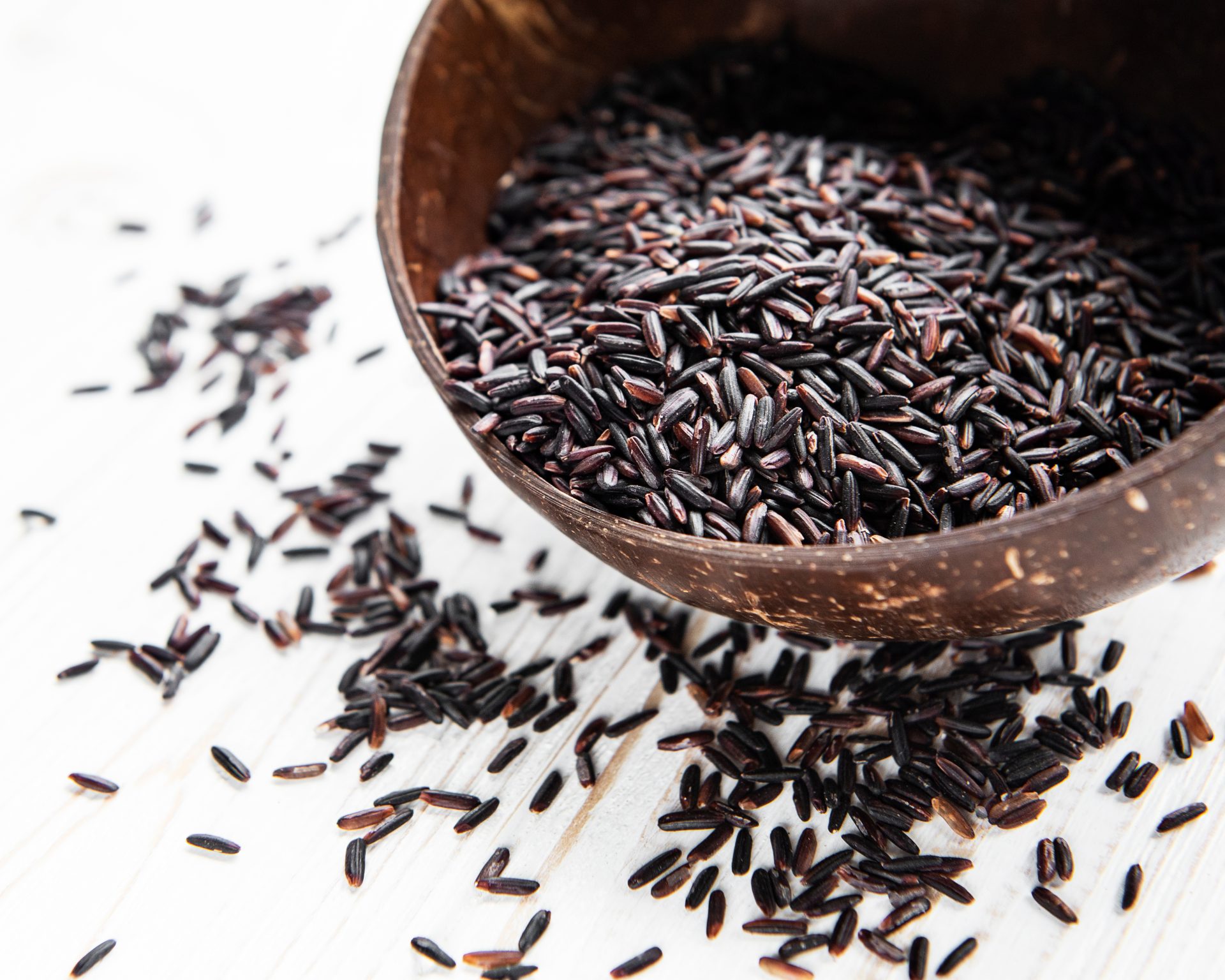 Closeup picture of a brown bowl full of dark wild rice grains, with some scattered below the bowl on a white surface.