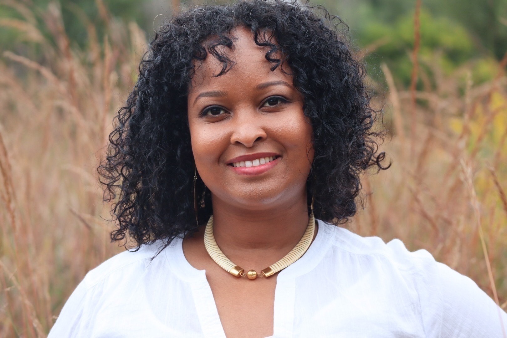 Naima Dhore headshot. Smiling woman stands in field of tall brown grass with greenery in distance