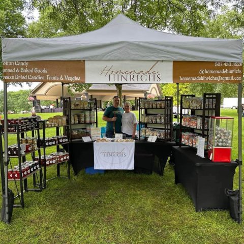 Picture of the Homemade Hinrichs farmers' market stand outside under a white tent canopy. Two people stand behind the table surrounded by homemade goods.