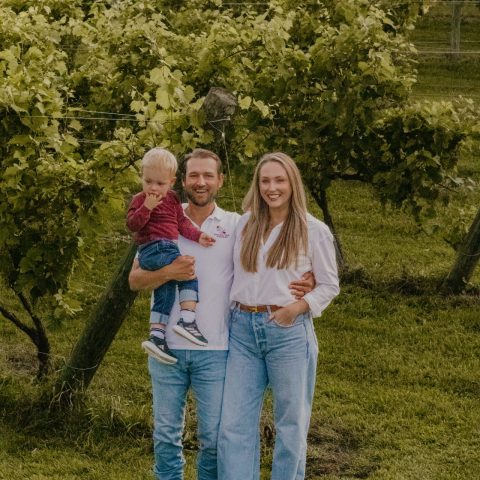 A smiling family of three stands in a sunny vineyard