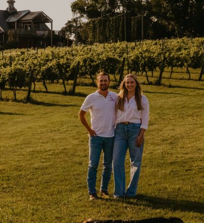 A happy couple stands in front of a vineyard