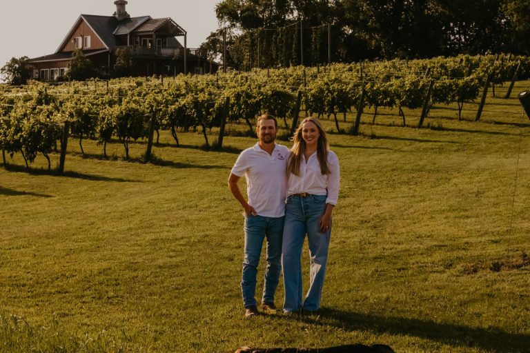 A happy couple stands in front of a vineyard