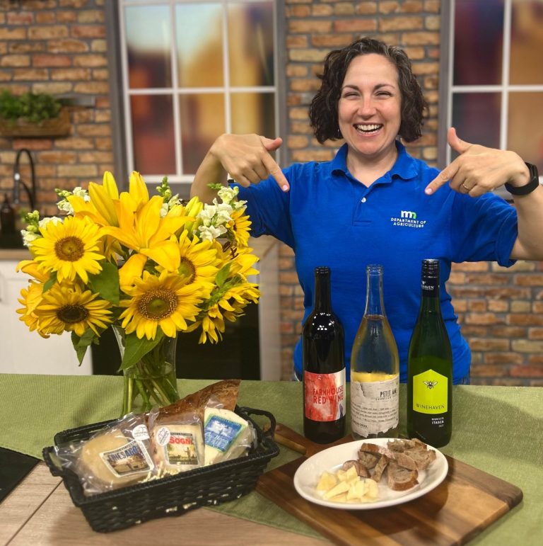 Rachel Wandrei points to counter with wine bottles, cheese, and bread. A vase of sunflowers sits off to the left