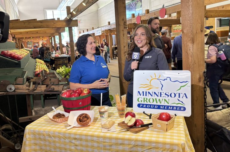 two women stand behind a table filled with apples and apples products. the minnesota grown stand sits in the foreground