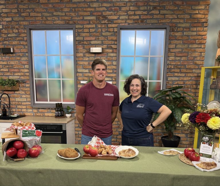 two people stand behind a counter smiling. apples and apple treats are displayed on the counter