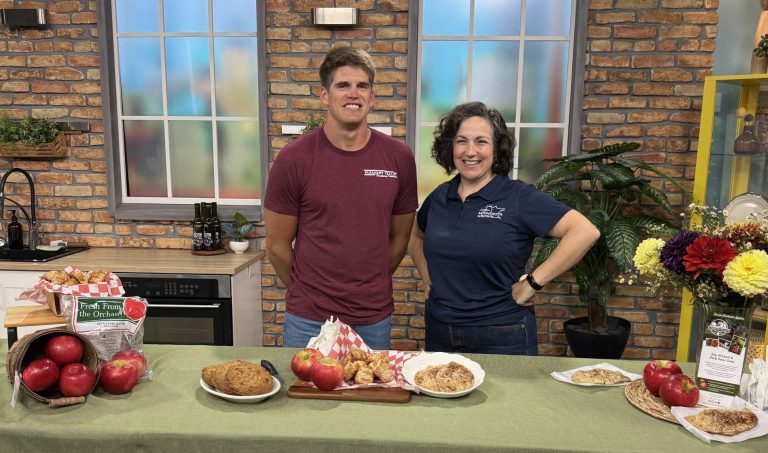 two people stand behind a counter smiling. apples and apple treats are displayed on the counter
