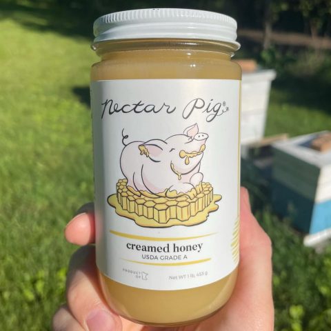 a single jar of nectar pig honey being held by a persons hands. beehive boxes are in the background