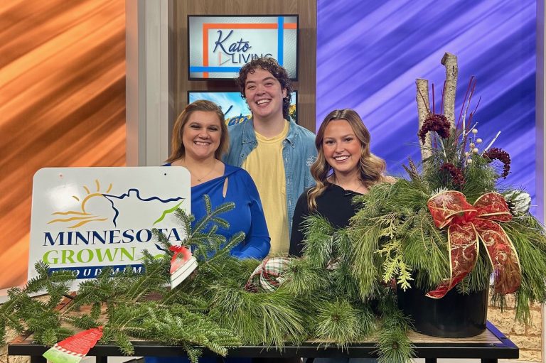 three people smile behind a table full of fir and pine greenery. a sign reads minnesota grown on the table, one reads kato living in the background