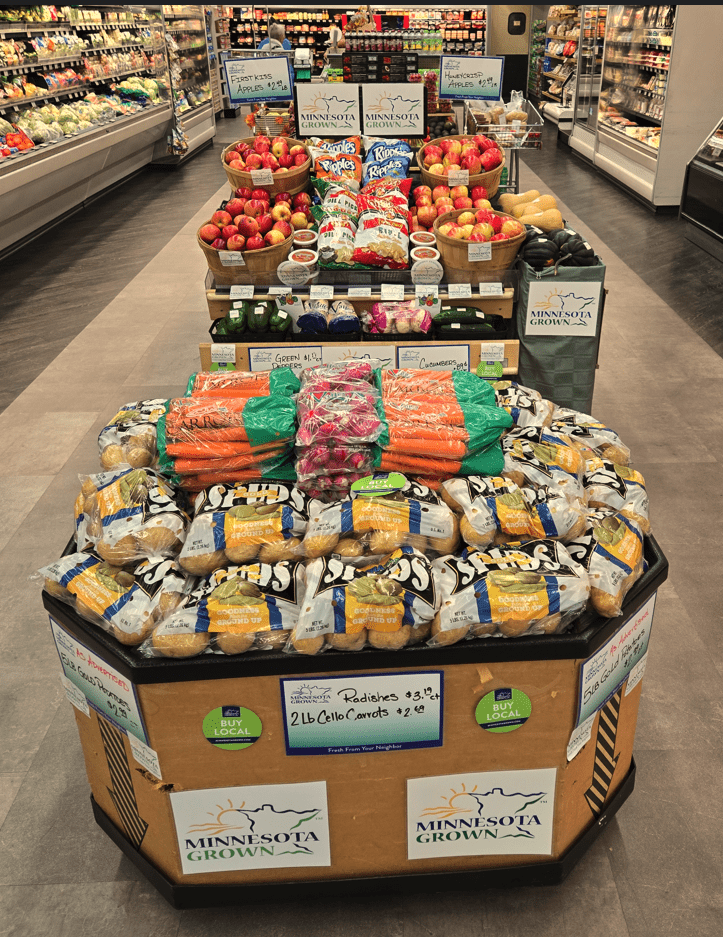 a grocery produce display in the middle of a wide aisle. features bags of potatos, carrots, and radishes. behind is another display full of apples and veggies. they all have signs that read "minnesota grown"