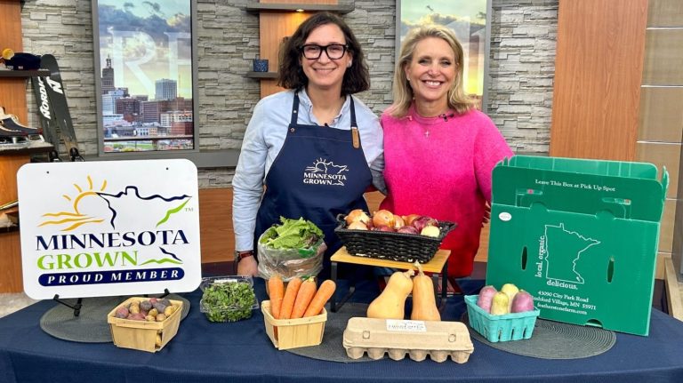 two women smile behind a counter full of fresh produce, a basket with Minnesota outline on it, and s a sign that reads Minnesota Grown