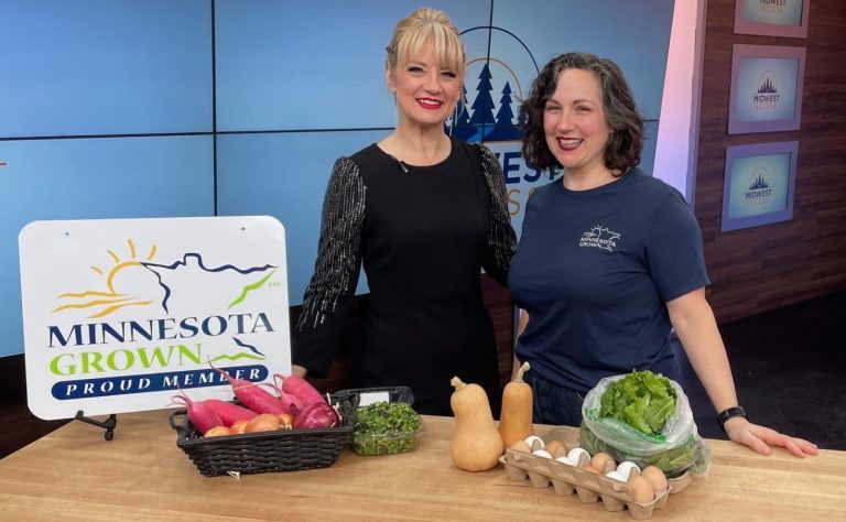 two women smile at a table full of fresh produce and a sign that reads "minnesota grown"