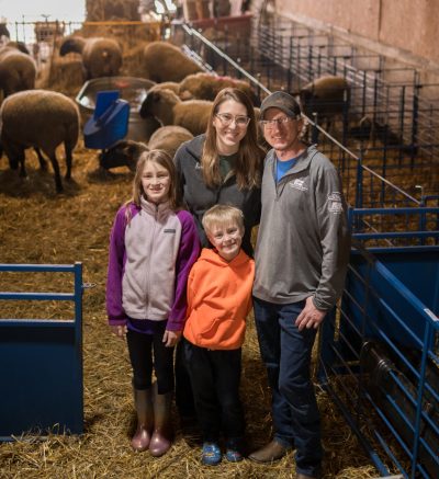 a family of four smile up at the camera, they are in a sheep barn surrounded by sheep