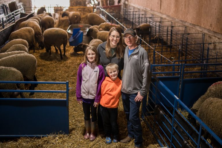 a family of four smile up at the camera, they are in a sheep barn surrounded by sheep