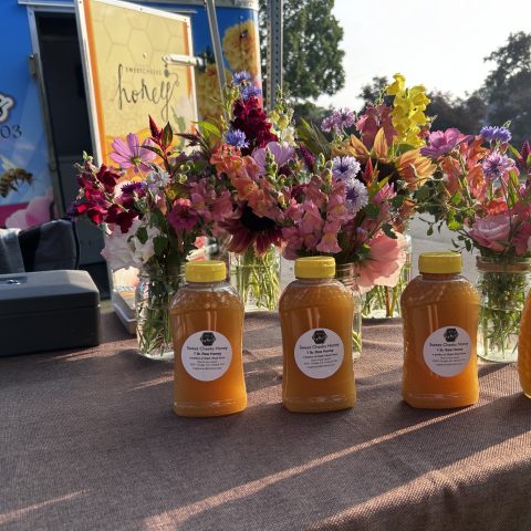 three bottoles of honey sit infront of three bouquets at a market stand