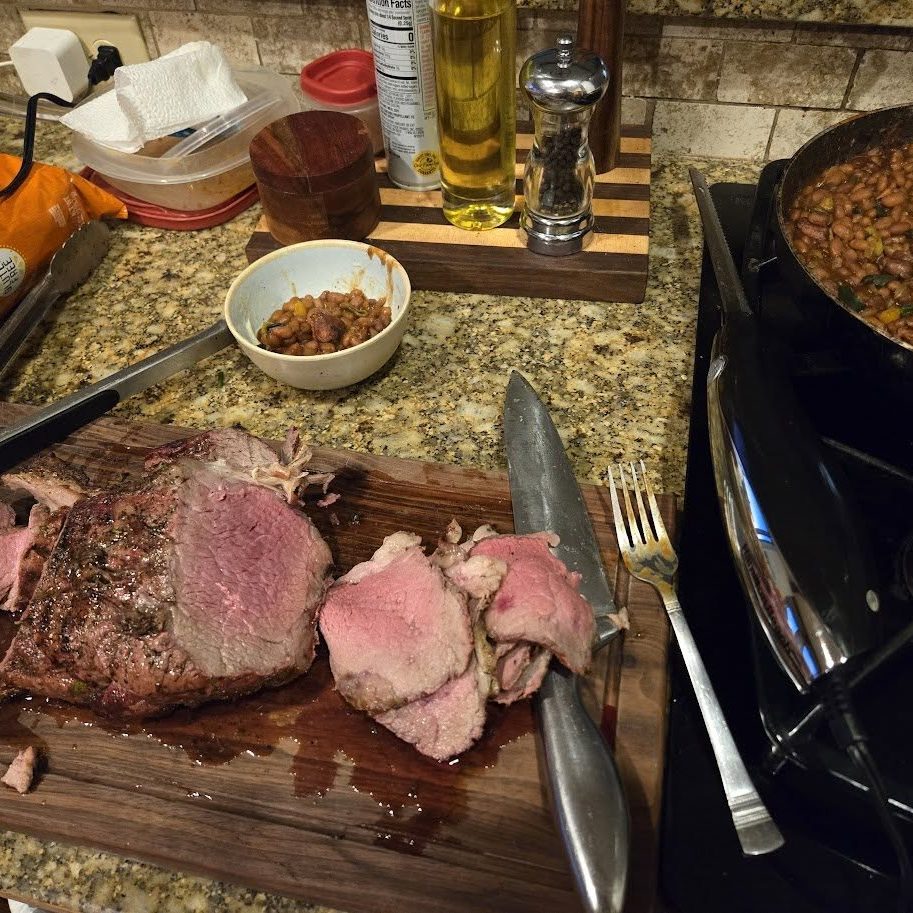 Roast beef on a counter. In the background is backed beans on the stove.