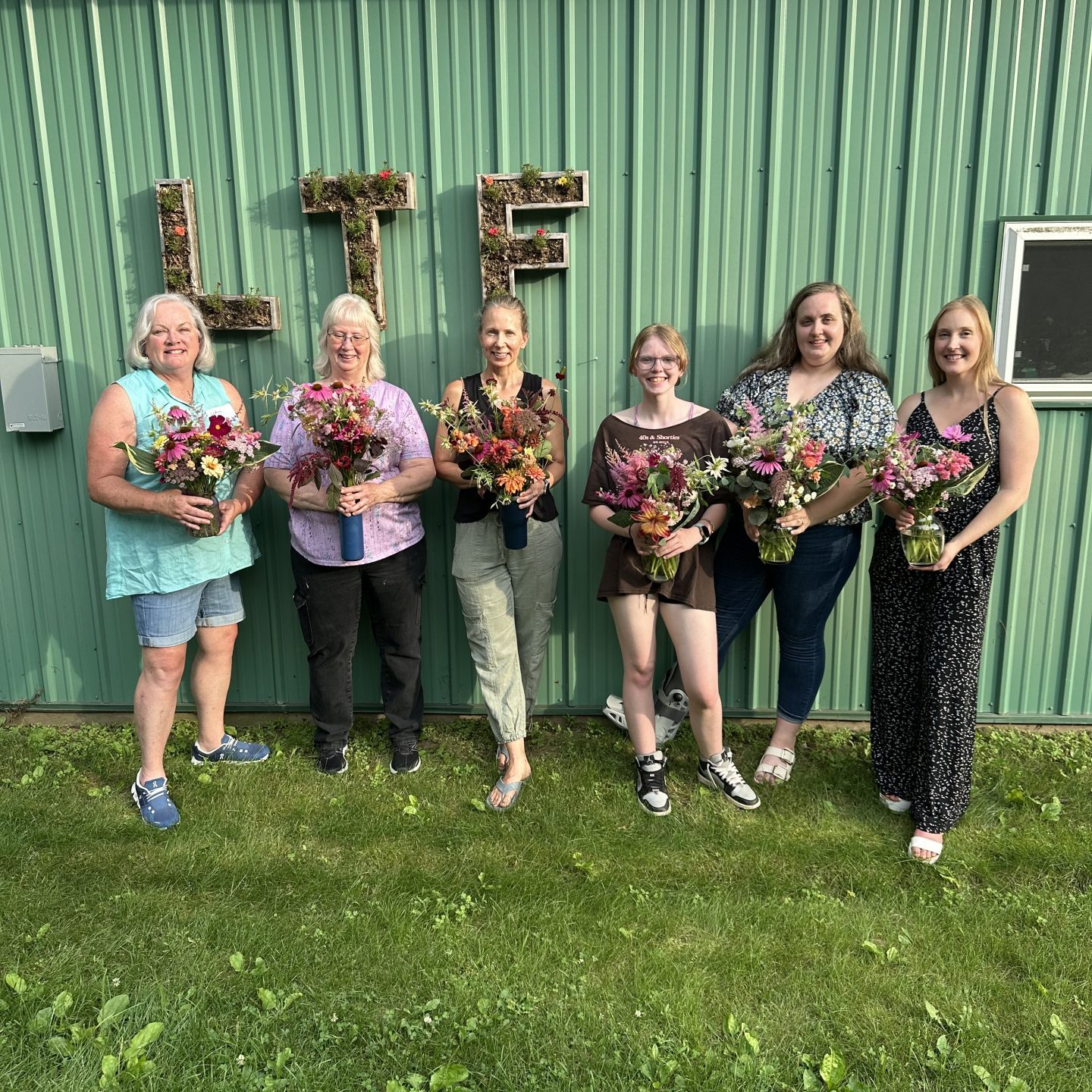 six women smiling and holding bouquets in front of a teal barn