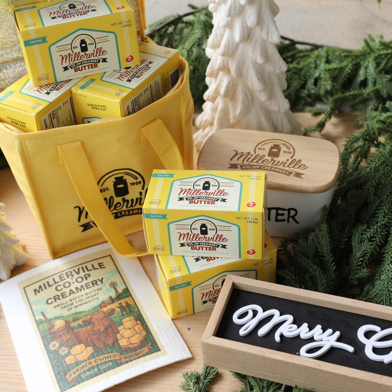 yellow tote filler with miller ville boxes, two pounds of pbutter stacked on one another, a white certamic butter dish, a swedish dishcloth and a merry christmas sign all surrounded by green boughs and white ceramic christmas tree décor